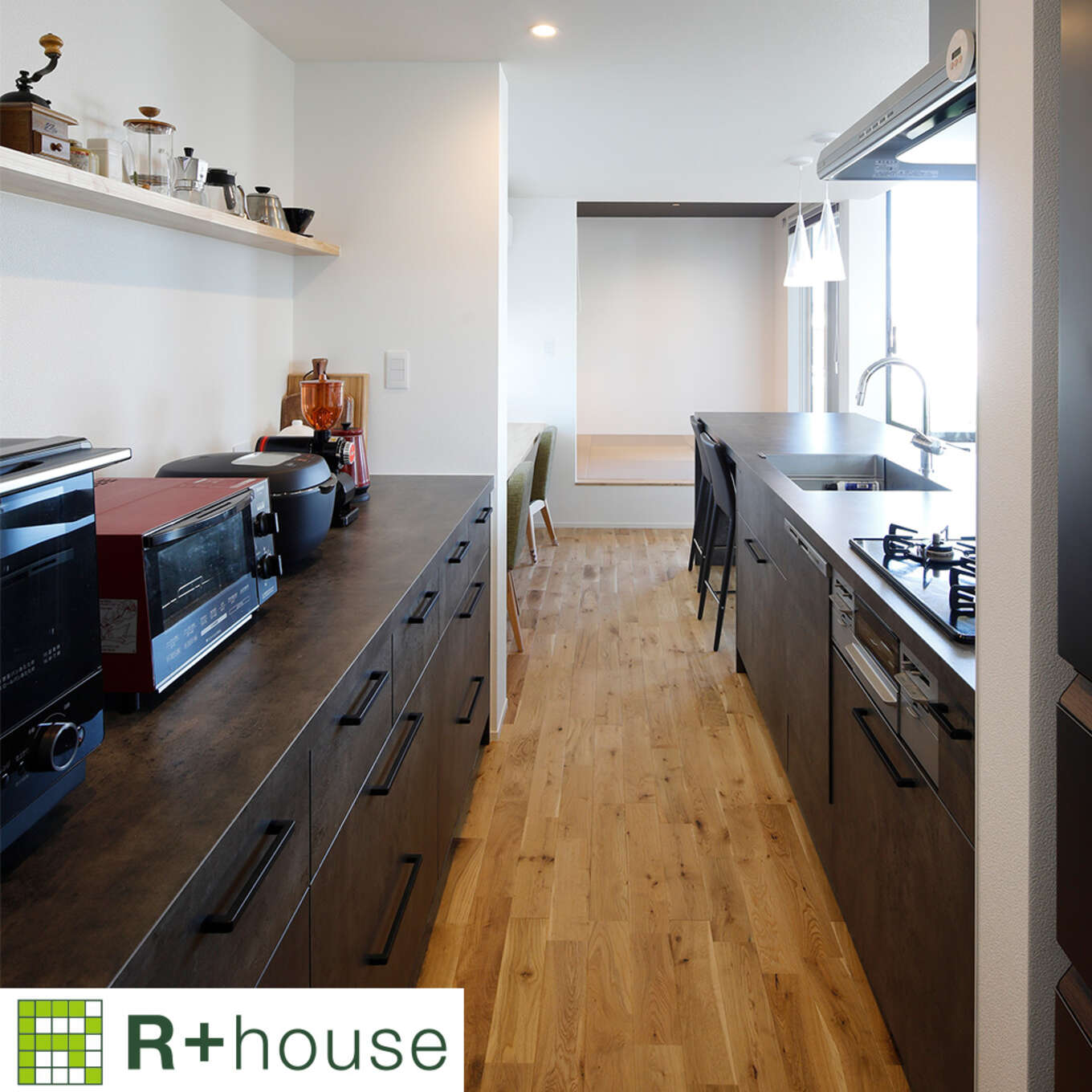 R+houseの物件のキッチンの写真です。左側は棚になっておりオーブンなど電化製品が置かれ、右側はコンロやシンク、ダイニングテーブルが並ぶ。