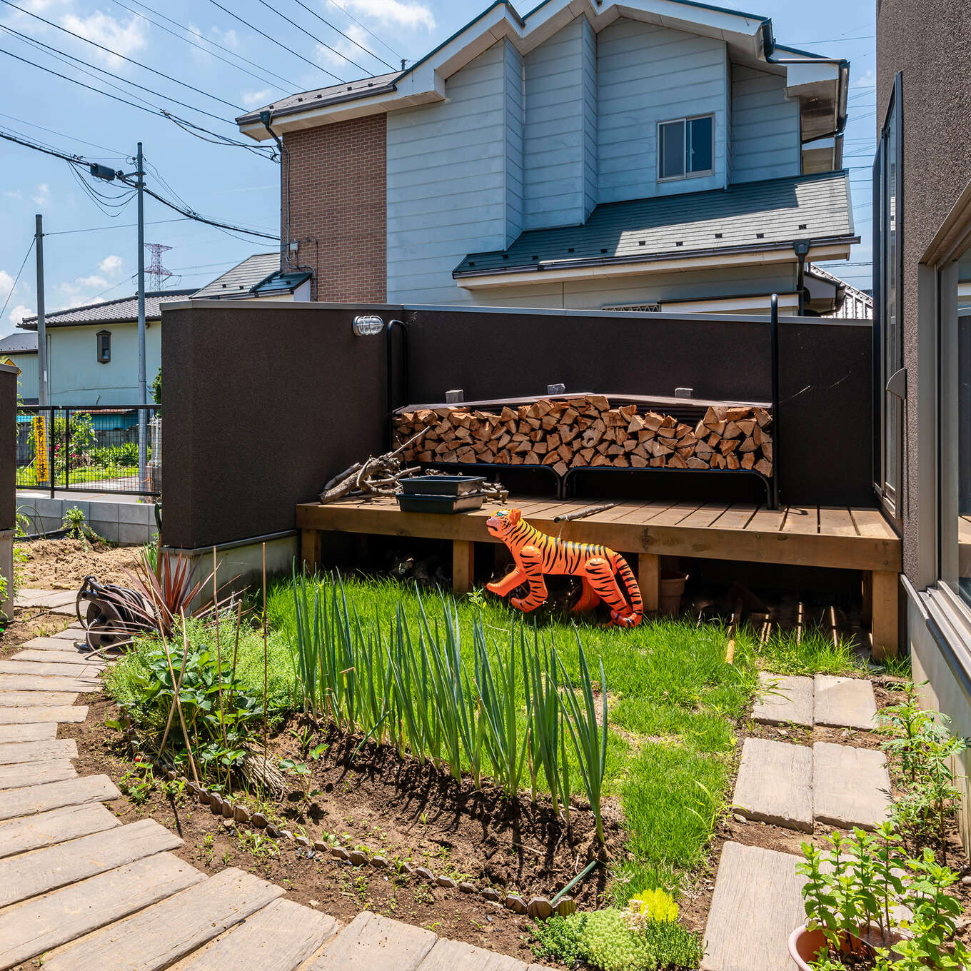 R+houseの物件の庭の写真です。奥にウッドデッキがあり、ストーブに使う薪が並んでいます。塀があるので外からは見えにくい。弧を描く園路の脇にネギなど野菜が植わっている