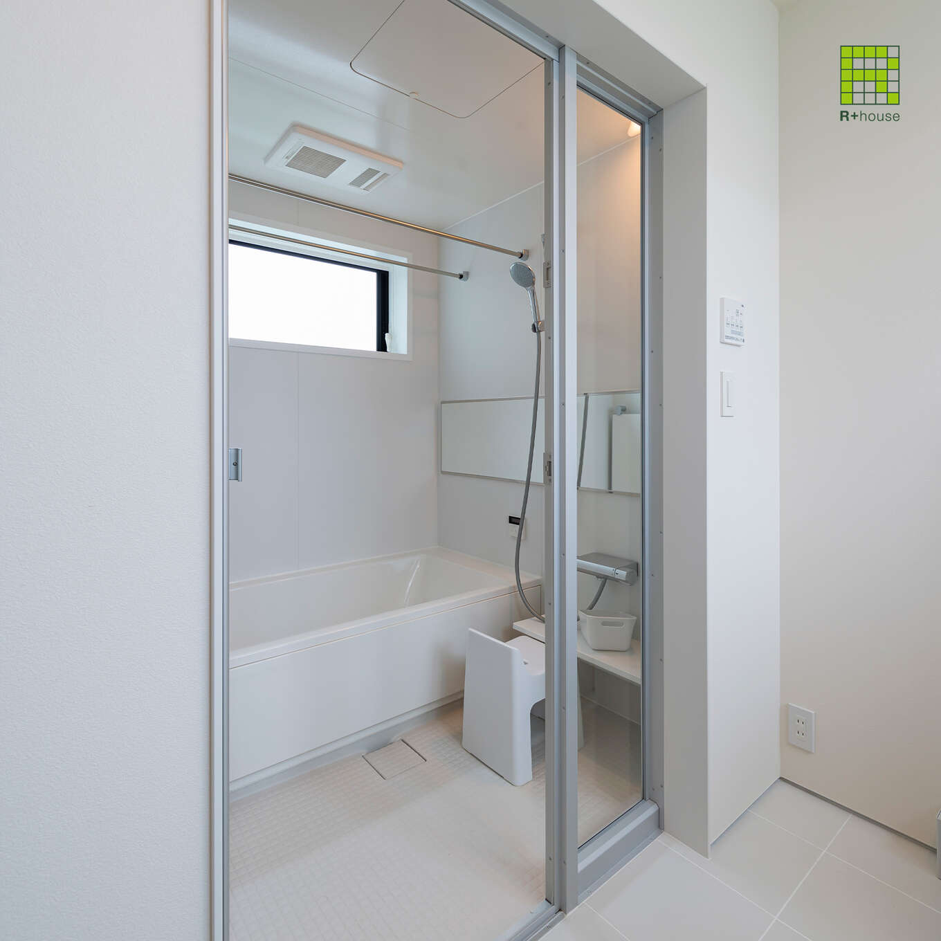 R+houseの物件の浴室の写真です。脱衣所からガラスの扉越しに真っ白な浴室が見える。
