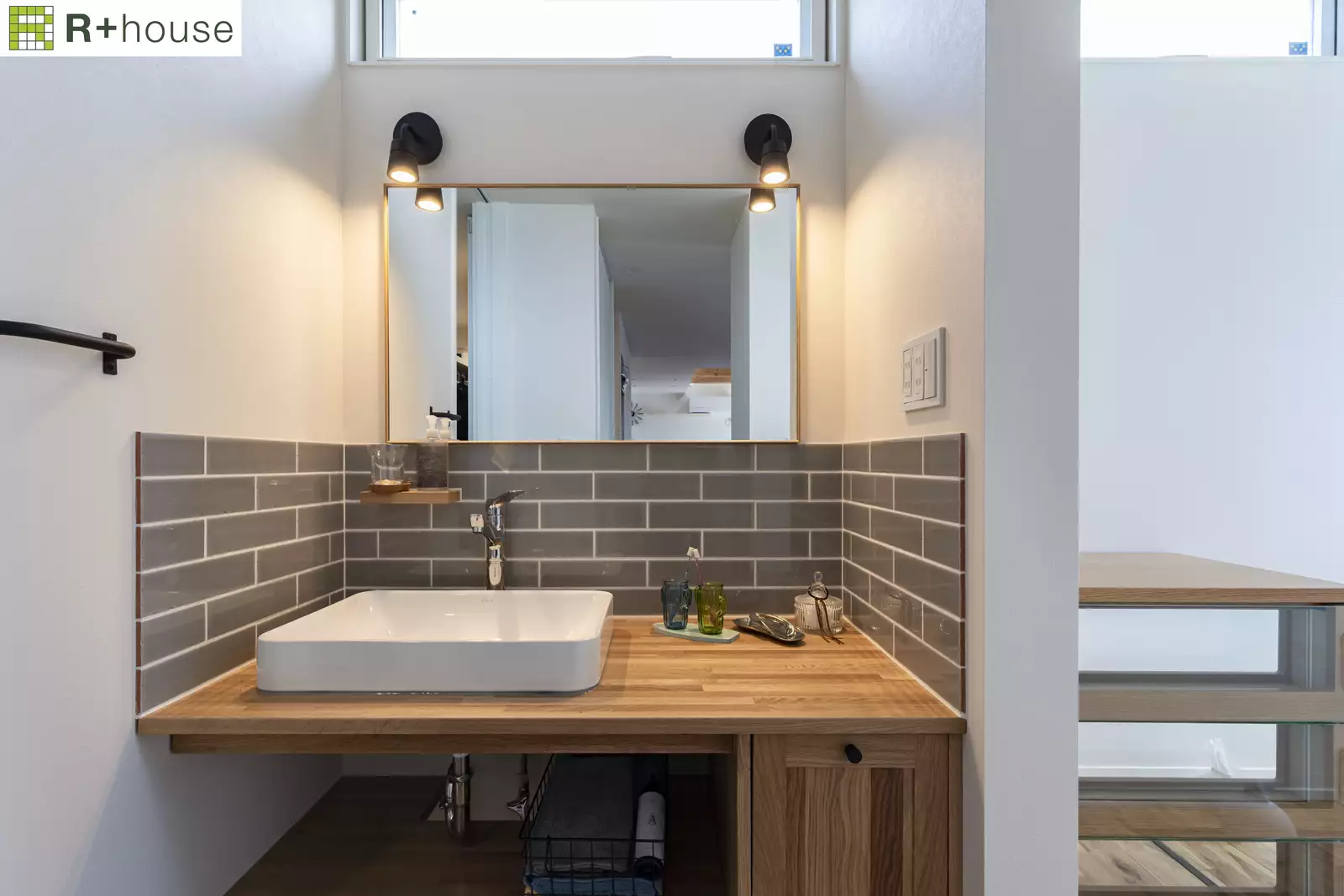 R+houseの物件の洗面所の写真です。白い壁の空間にグレーのタイルを張り、木のカウンターの上に白いボウルの洗面台を設けています。