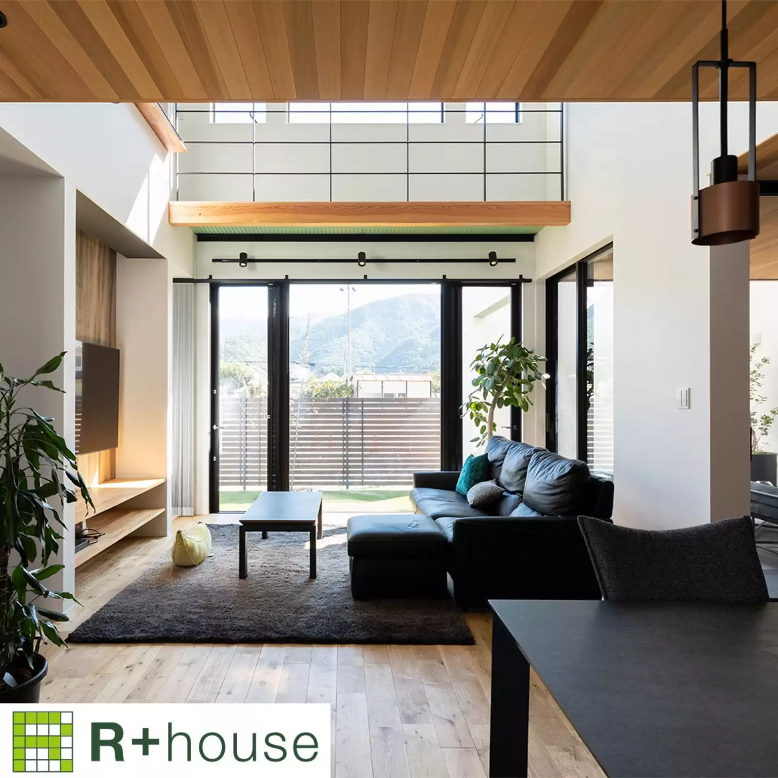 R+houseの物件のリビングの写真です。左側に壁かけテレビ、右側にソファー。奥にある窓からは芝の庭が見え、吹き抜け越しには山々を遠望できる。