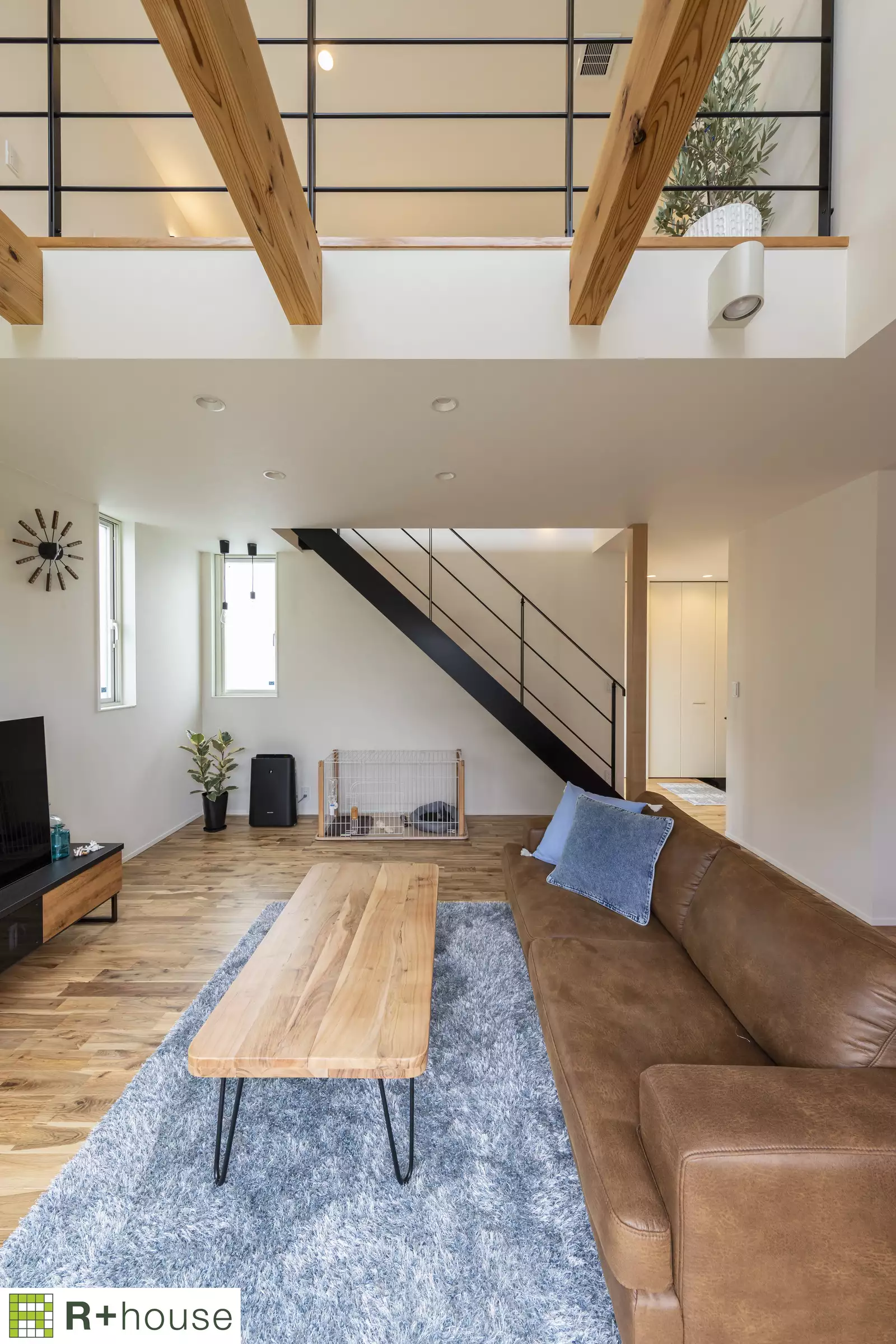 R+houseの物件のリビング写真です。ソファにローテーブル、グレーの絨毯、奥に二階へ続く階段が見えます。