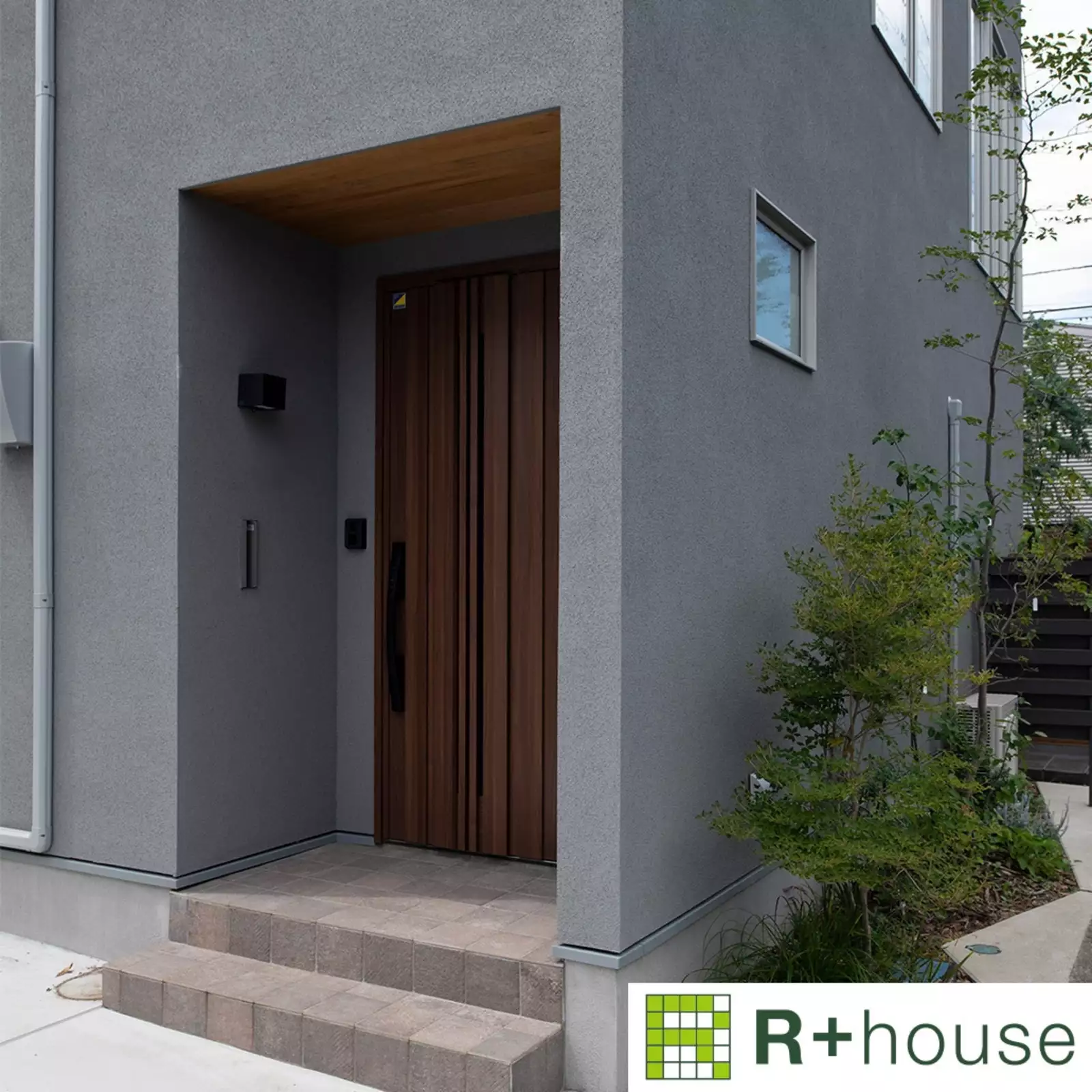 R+houseの物件の玄関の写真です。壁はグレー、濃いブラウンのおしゃれなドア。天井は木目調の明るいブラウン