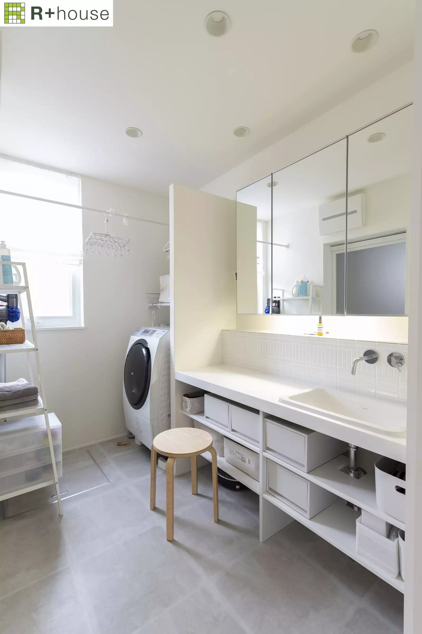 R+houseの物件の洗面所の写真です。グレーのタイルに白い洗面台と白のドラム式洗濯機があります。