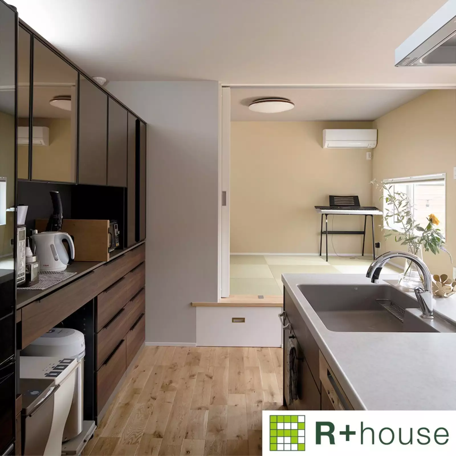 R+houseの物件のキッチンの写真です。右手にアイランド型のキッチンがあり、左手には落ち着いたダークブラウンの棚に家電が並ぶ