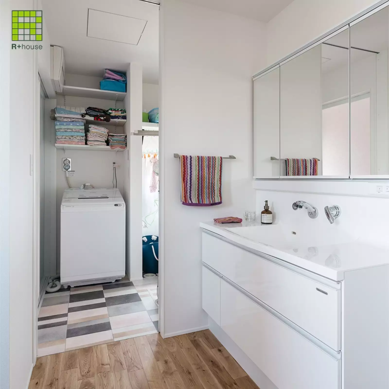 R+houseの物件の洗面台の写真です。白い大き目の洗面台は使い勝手がよさそう。奥には洗濯スペースと脱衣室がある。