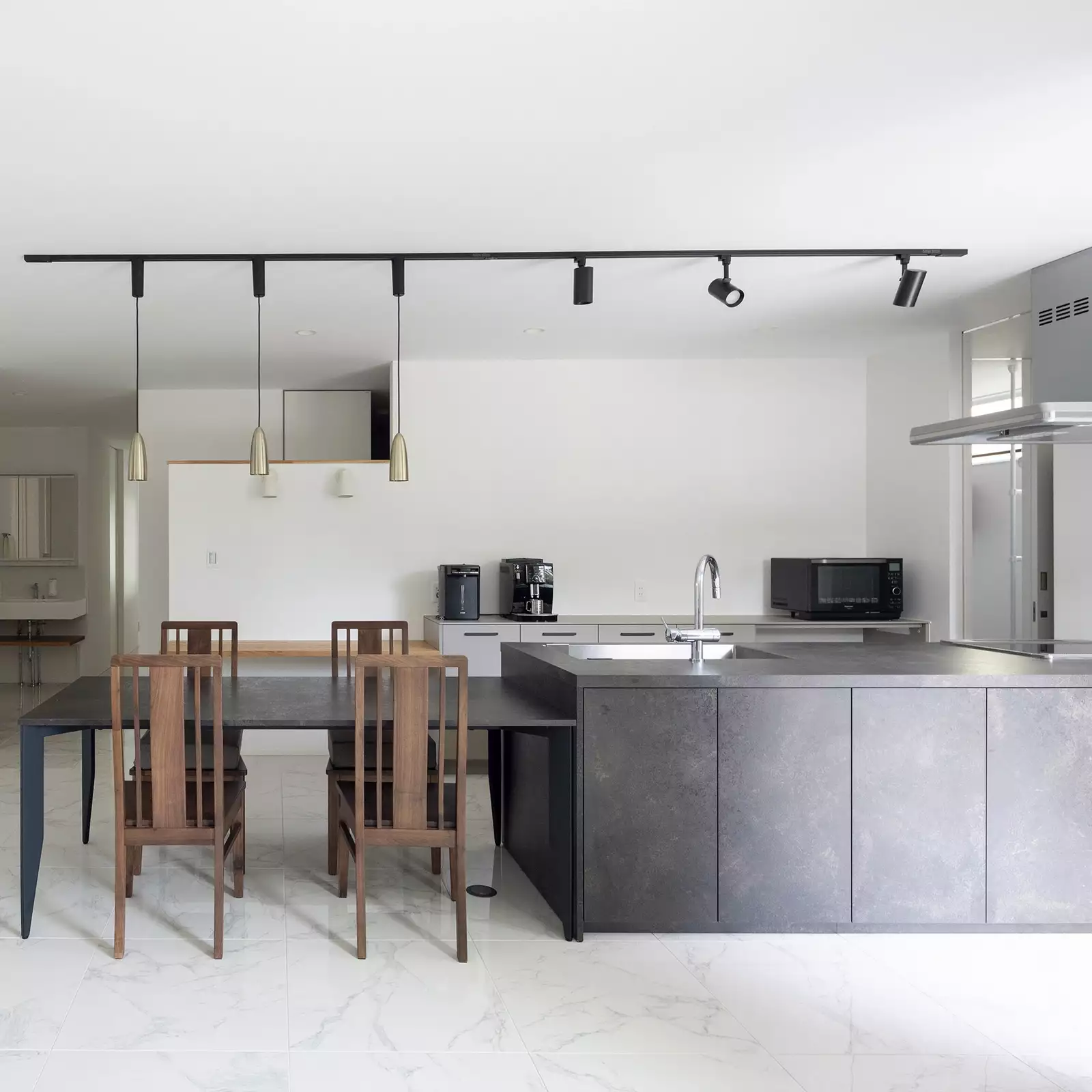R+houseの物件のキッチンの写真。濃いグレーで統一されたアイランドキッチンとダイニングテーブル。白いタイルの床と相まっておしゃれ。
