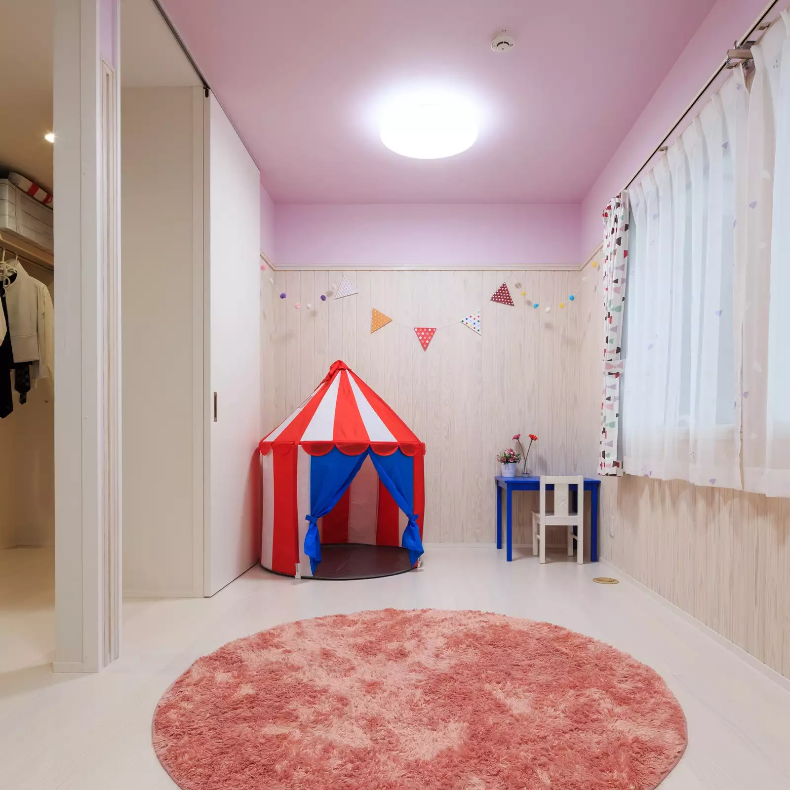 R+houseの物件の子ども部屋の写真です。白い床、白い木の腰板。天井と壁の上の方はピンクの壁紙でかわいい。右側に窓、左側にはドア。ドアの向こうにファミリークローゼット。
