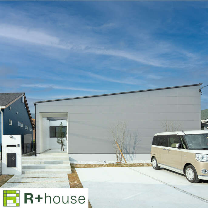 R+houseの物件の写真です。広々とした駐車場の奥にある明るいグレーの平屋です。