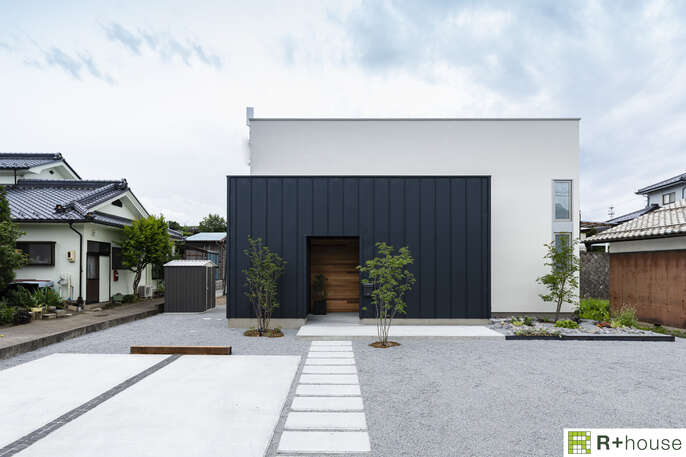 R+houseの住宅の外観写真です。白と黒の建物の前に2台分の駐車スペースとアプローチ、植栽があります。