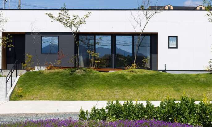 R+houseの物件の写真。白い平屋の家。駐車スペースとは高低差があり、芝生や木々が植えられている。