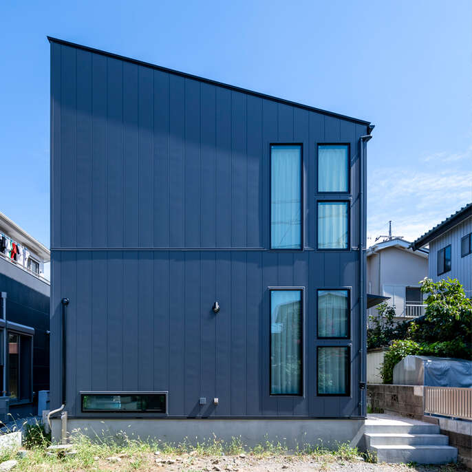 R+houseの物件の外観の写真です。紺色の外壁。左側の屋根が高く、右側の屋根が低い形状。右側の壁には窓が並ぶ